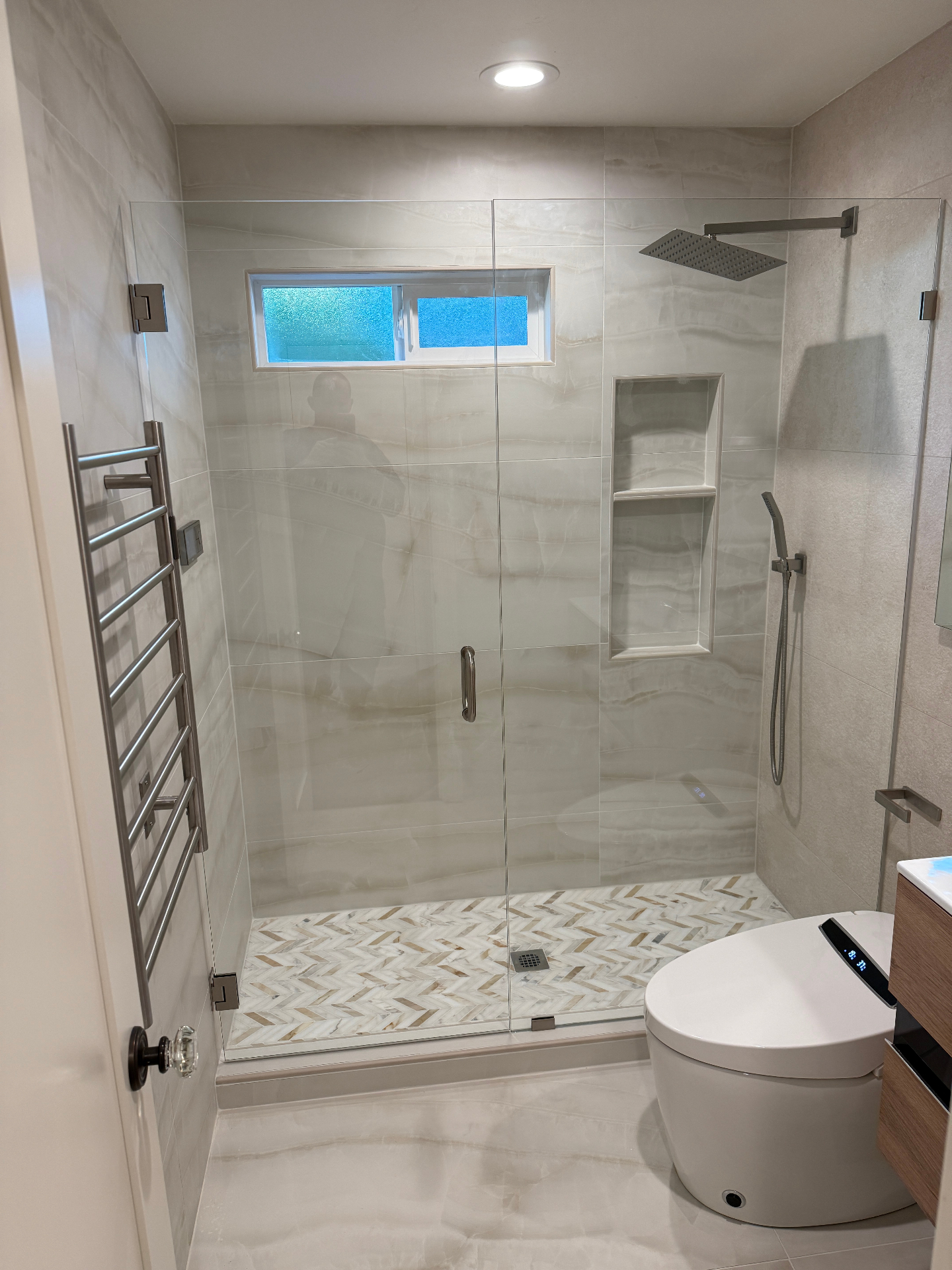 Wide view of finished beige bathroom showing frameless glass shower, niche, and herringbone floor
